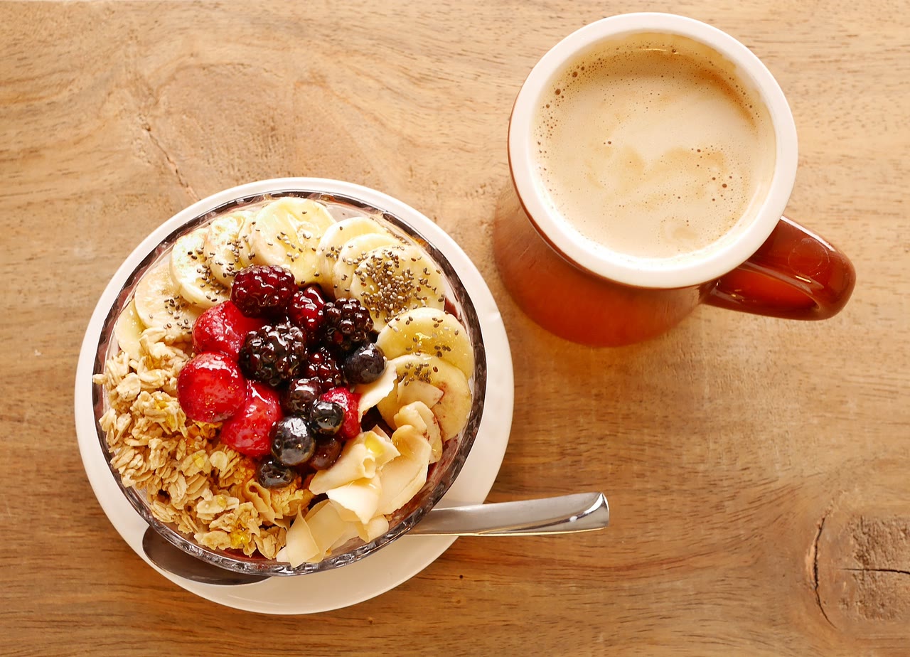Bowl de granola con banana, fresas, zarzamoras, moras azules y coco, junto a una taza de café